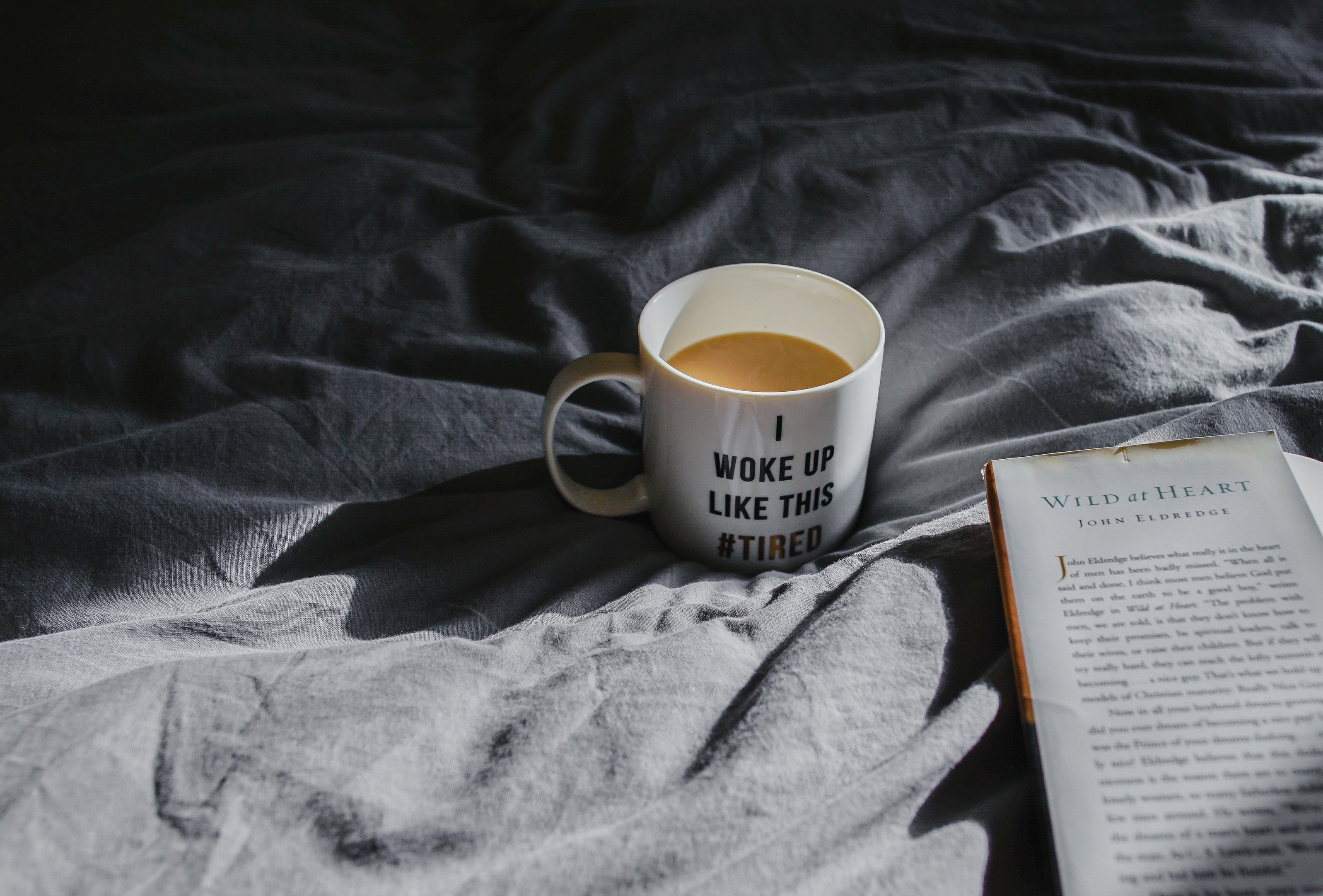 White mug of coffee reading I WOKE UP LIKE THIS #TIRED on rumpled gray sheets beside an open book in morning light