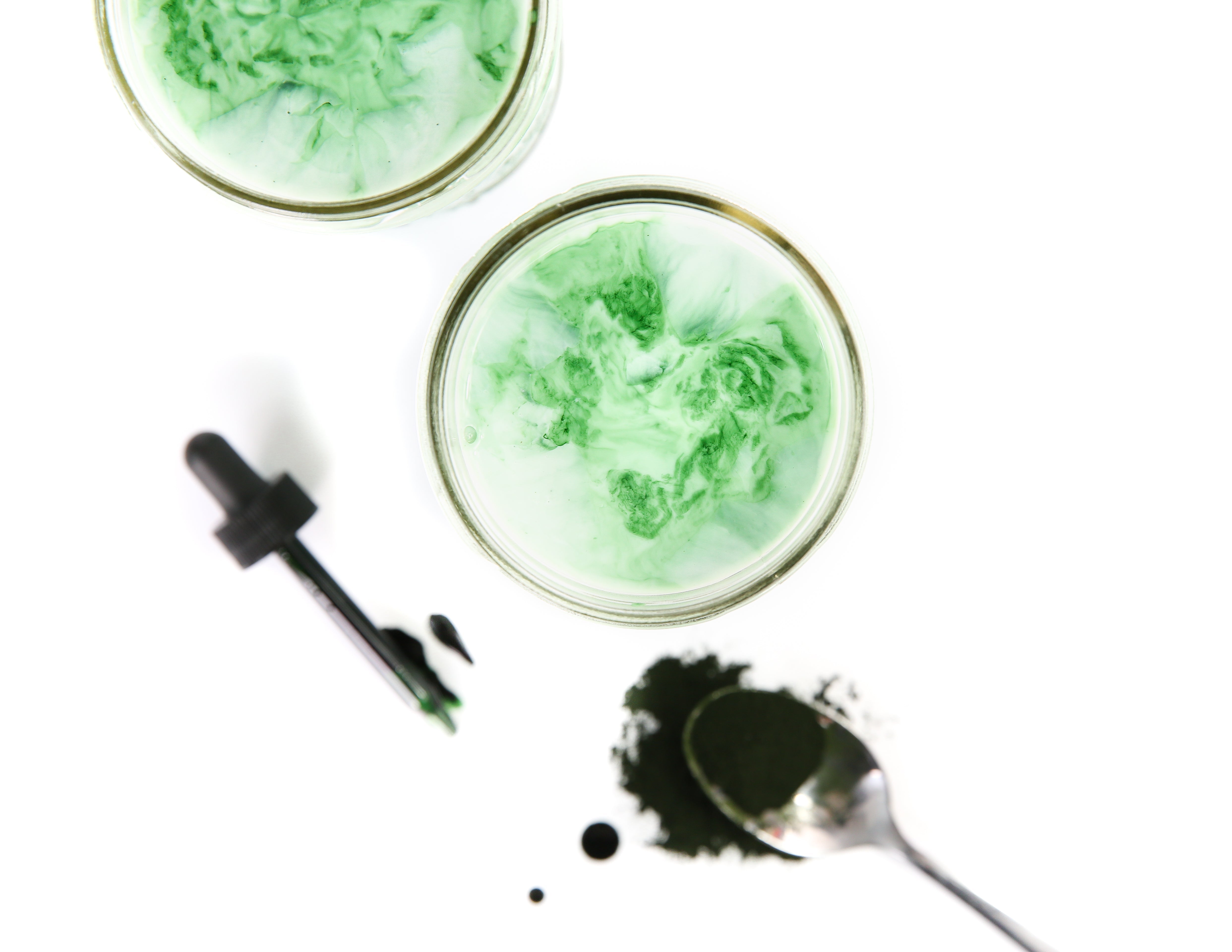 Top-down view of two green recovery tonics in glasses with a dropper and a spoon of green powder on a white background