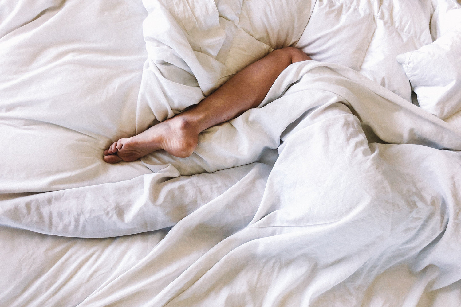 Top-down view of a person lying in bed with one bare leg exposed from rumpled white sheets