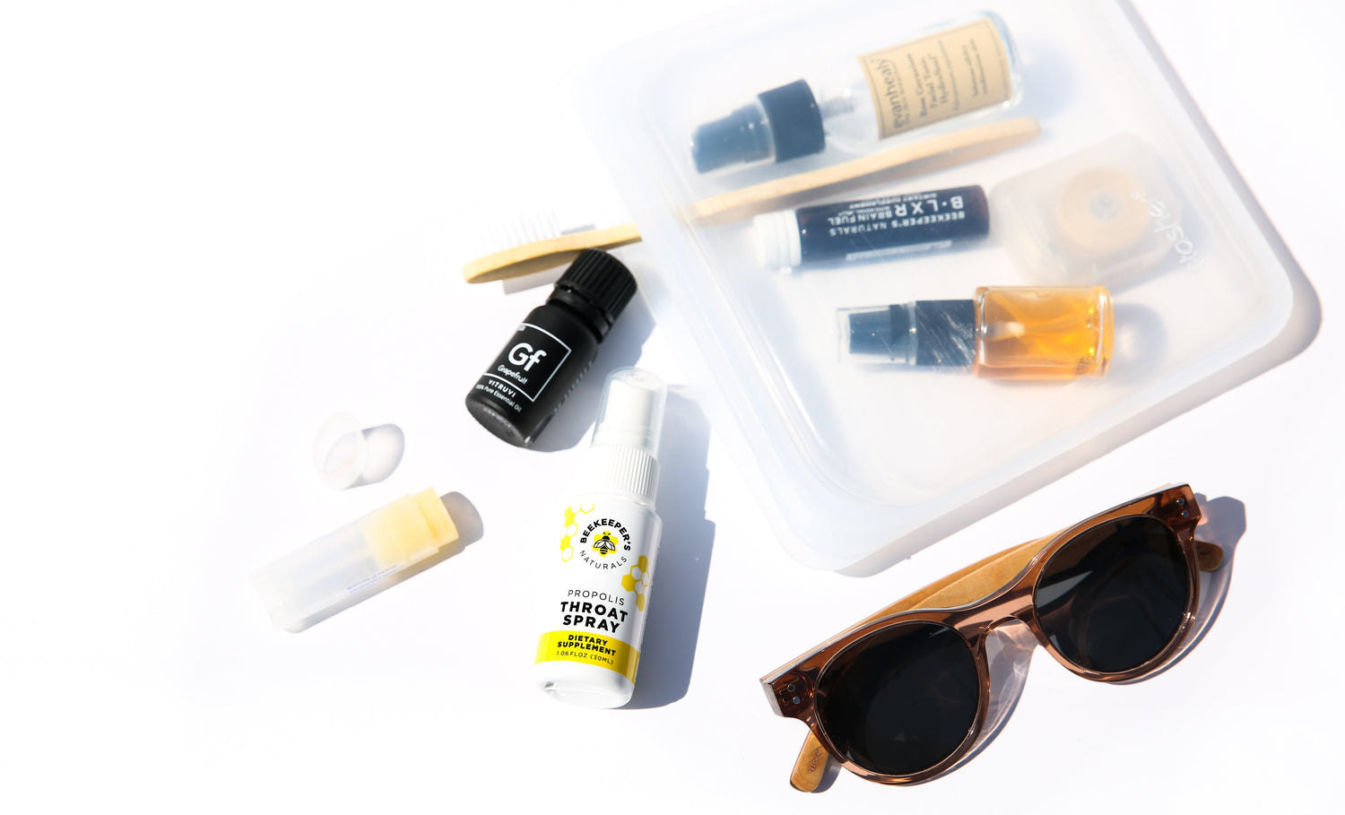 Flat lay of travel essentials: Beekeeper's Naturals Propolis Throat Spray, lip balm, small travel bottles, sunglasses, and clear travel case on a white background