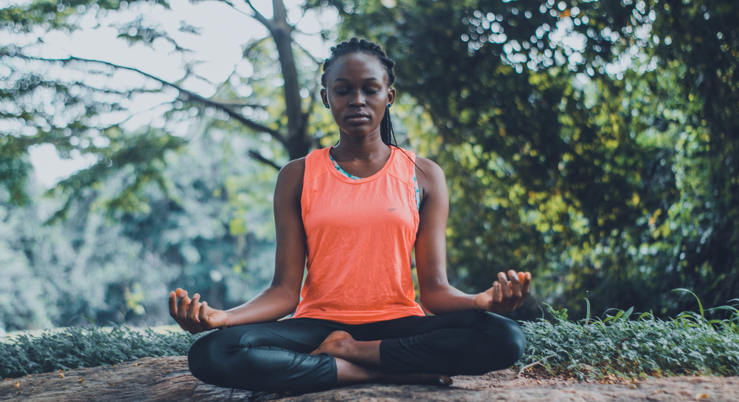 Woman meditating cross-legged on a log in a leafy park, wearing an orange tank top and black leggings