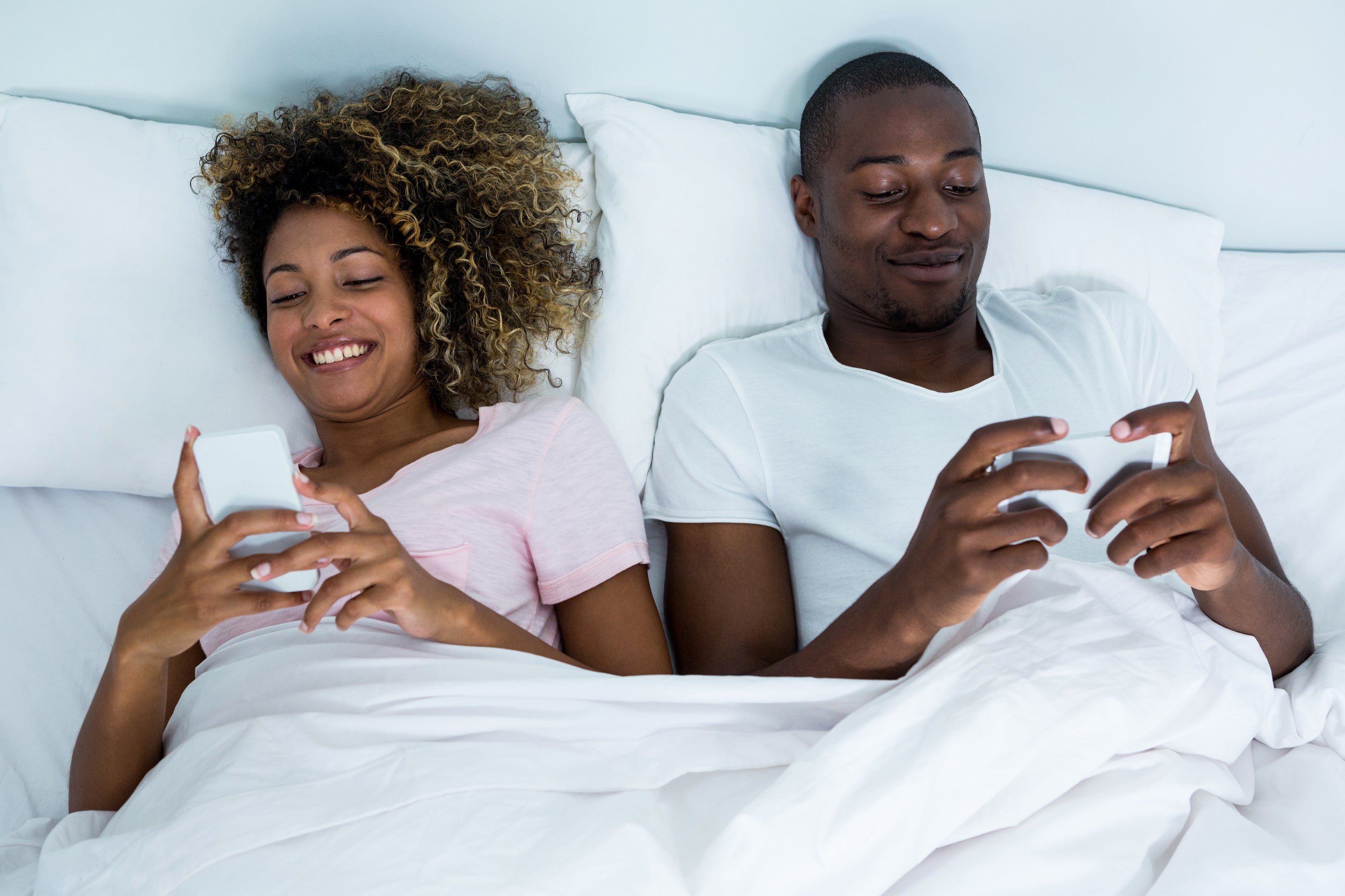 Couple lying in bed using smartphones, screens glowing as they scroll before sleep