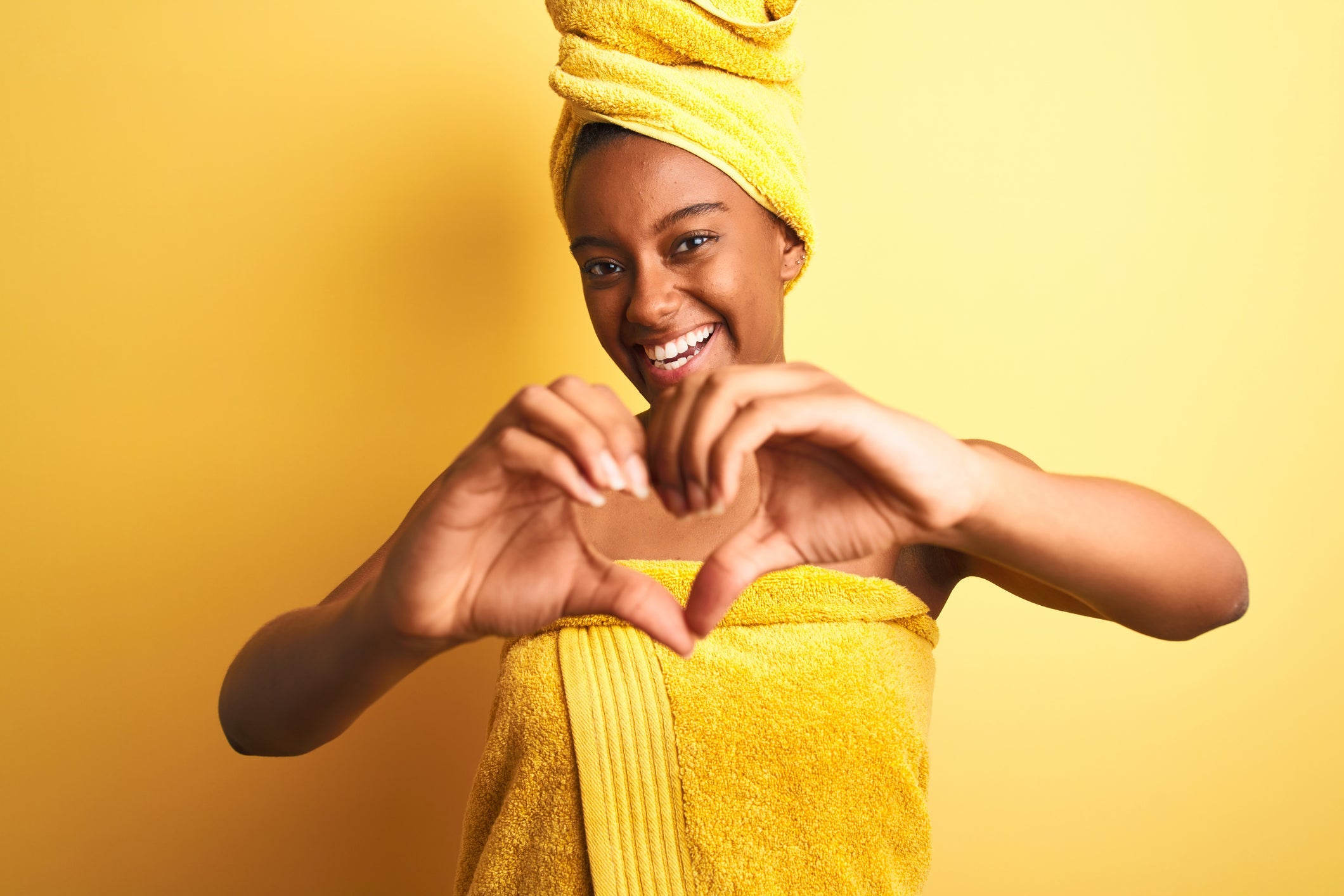 Young woman wrapped in a yellow towel and matching hair turban smiling and forming a heart with her hands against a yellow background