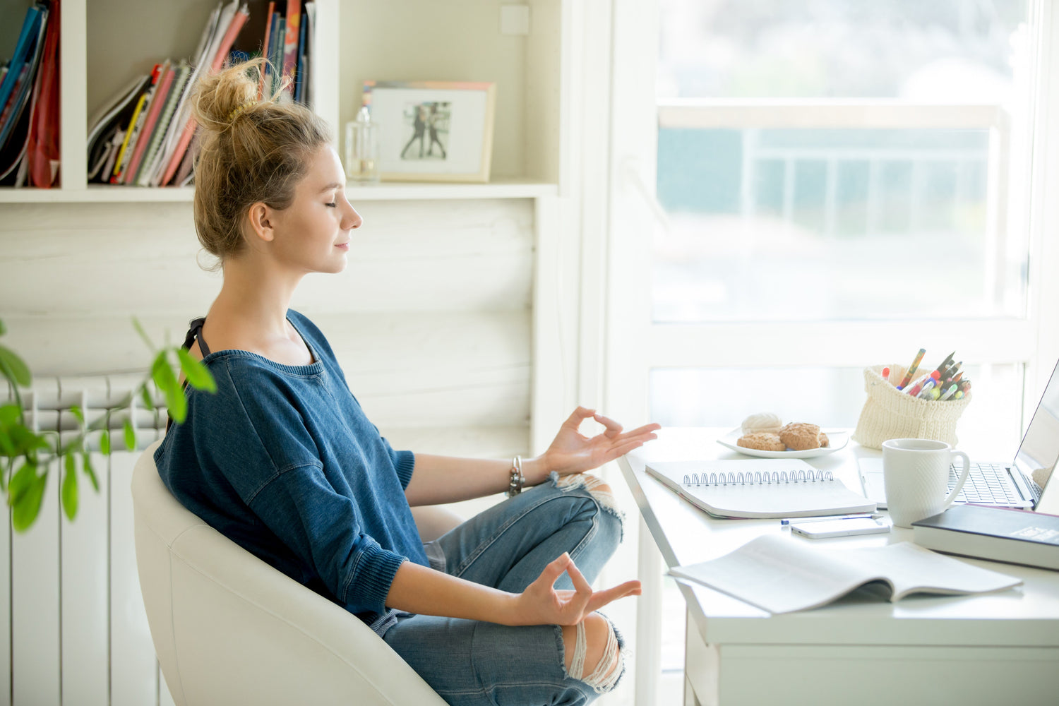 Woman meditating at a sunlit home desk during the workday with a laptop, notebook, mug, and snacks nearby
