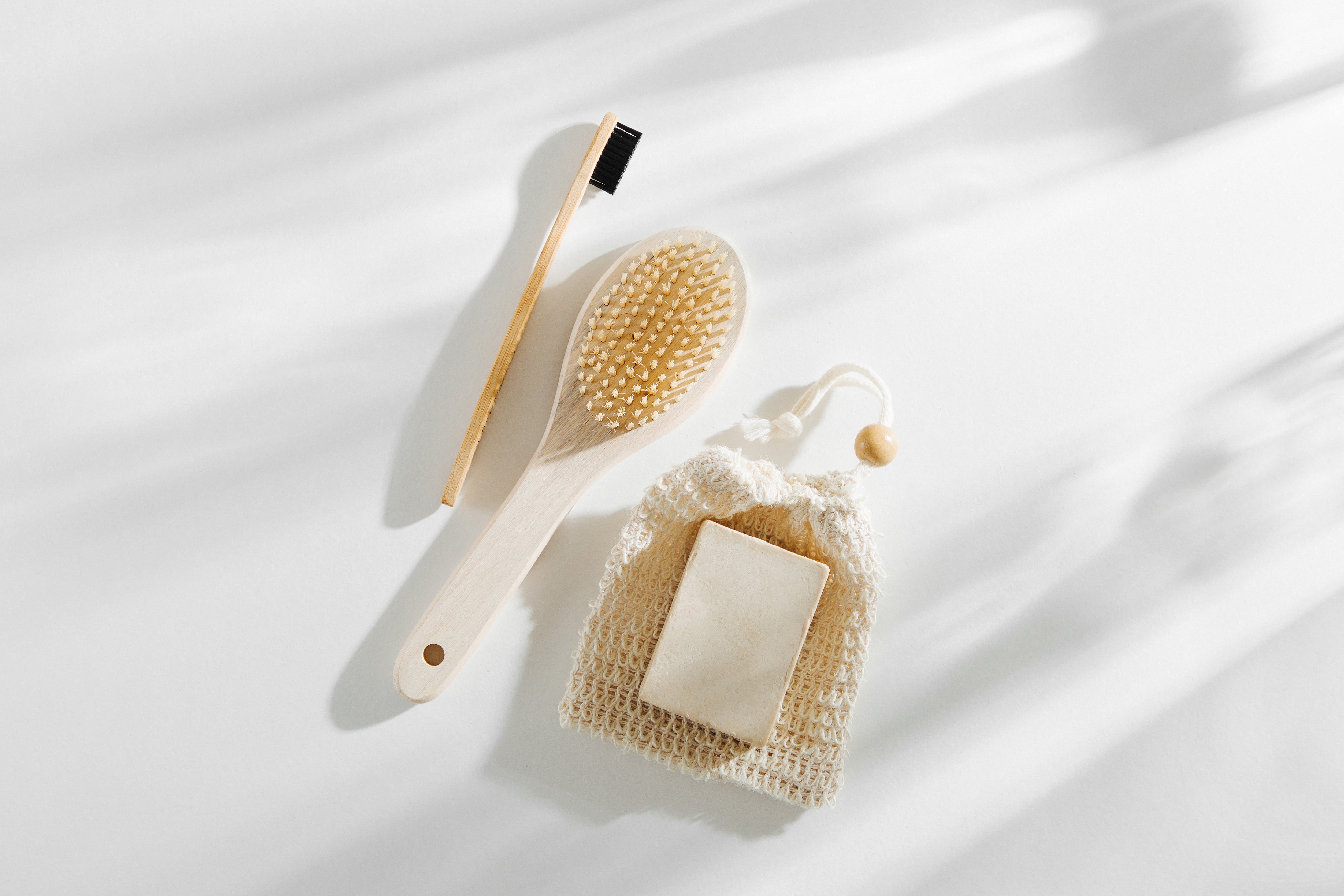 Wooden toothbrush, dry body brush, and soap bar in a mesh pouch arranged on a sunlit white surface