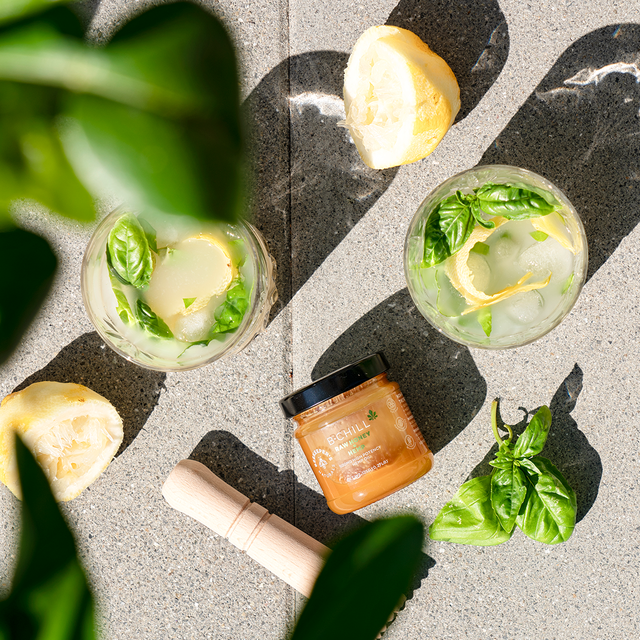 Two basil lemon-aid drinks with ice beside a B.Chill honey jar, lemon halves, fresh basil leaves and a wooden muddler on sunlit concrete