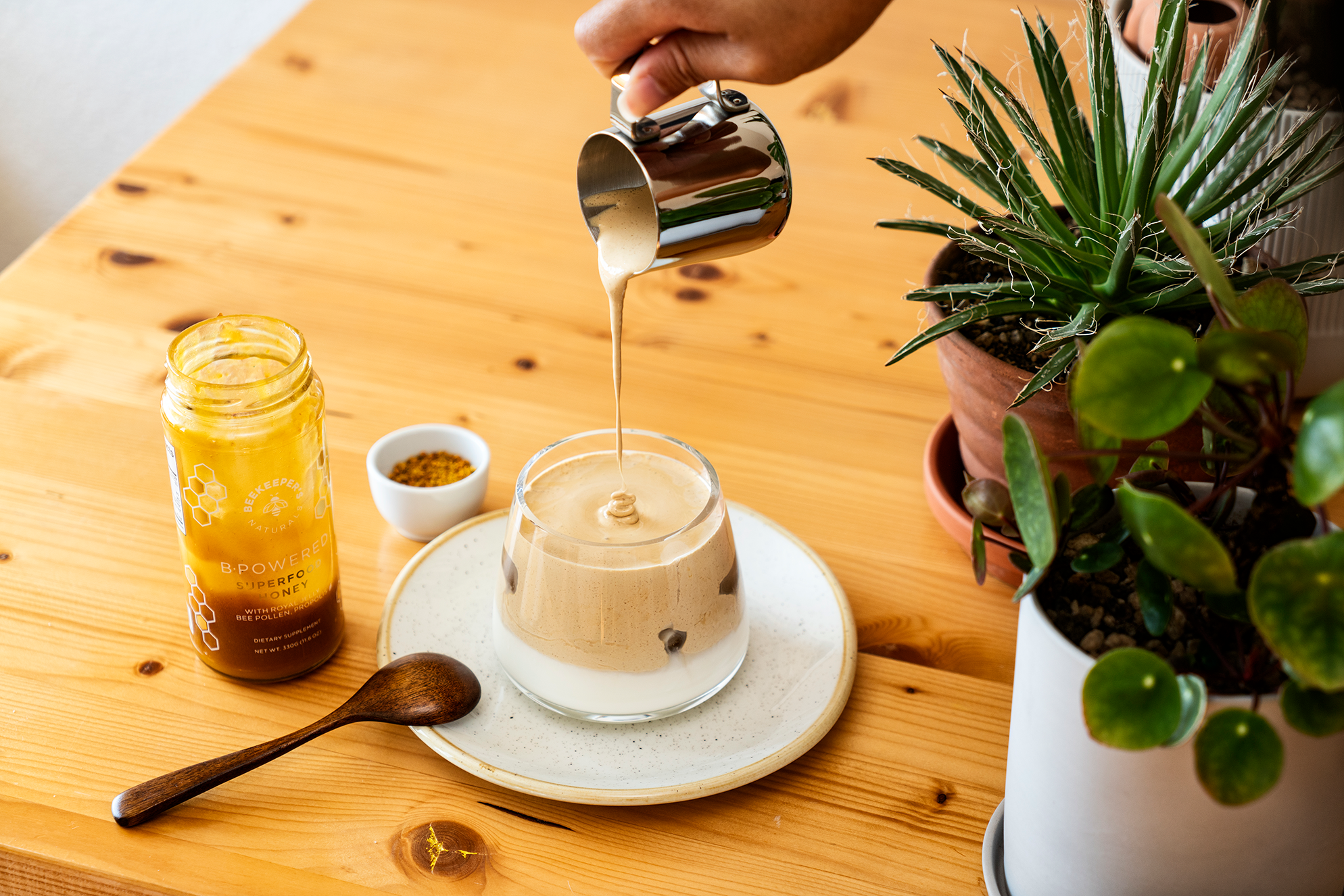 Whipped dalgona coffee being poured over milk in a glass on a wooden table with a jar of B Powered honey and potted plants