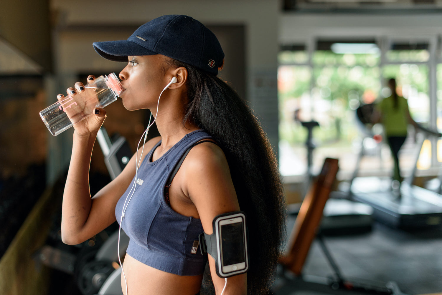 Woman drinking from a clear sports bottle in a gym, wearing a navy crop top, baseball cap, earbuds, and a phone armband