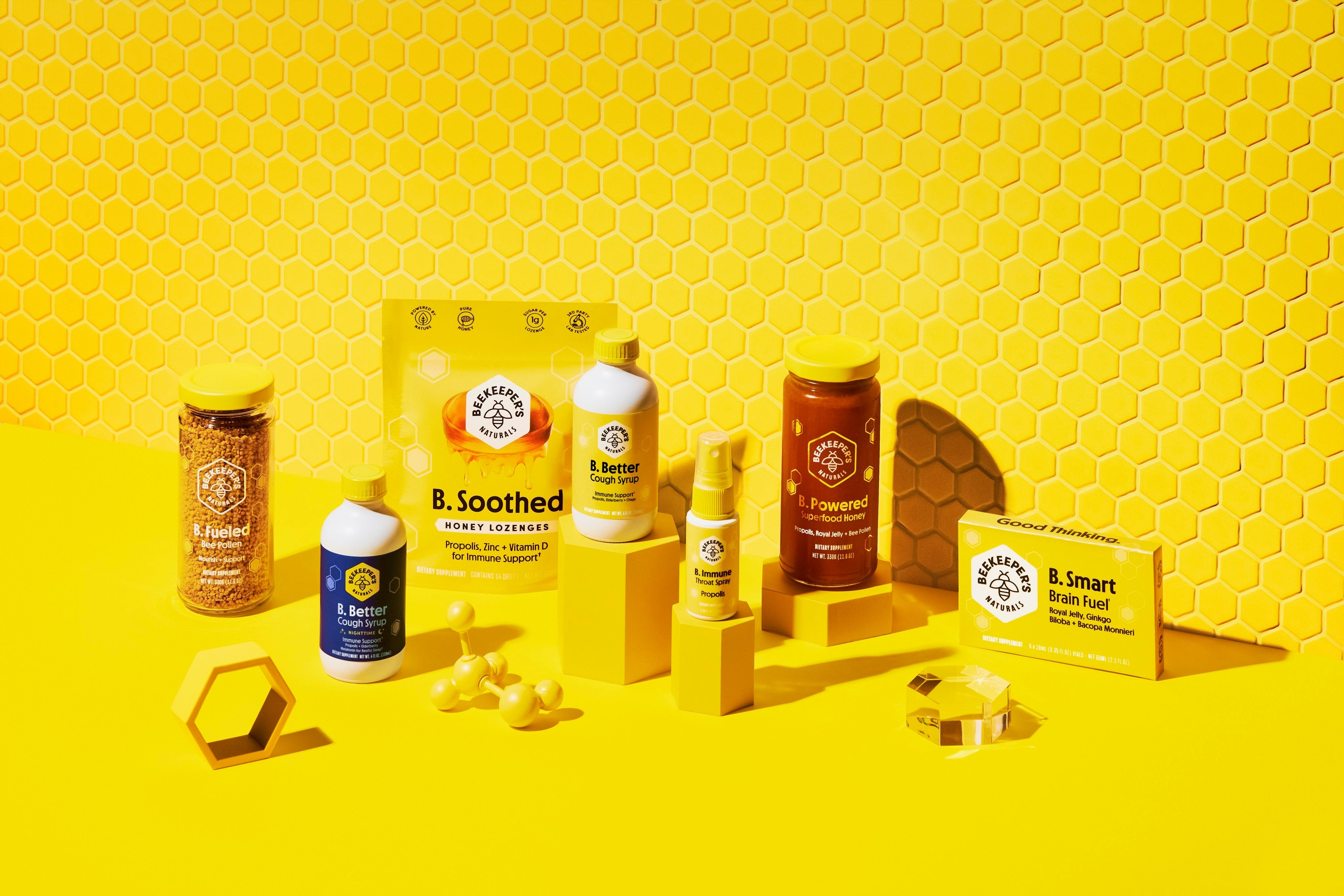 Front view of assorted Beekeeper's Naturals products (B. Soothed, B. Better, B. Powered, B. Smart, B. Fueled) displayed on a bright yellow honeycomb background
