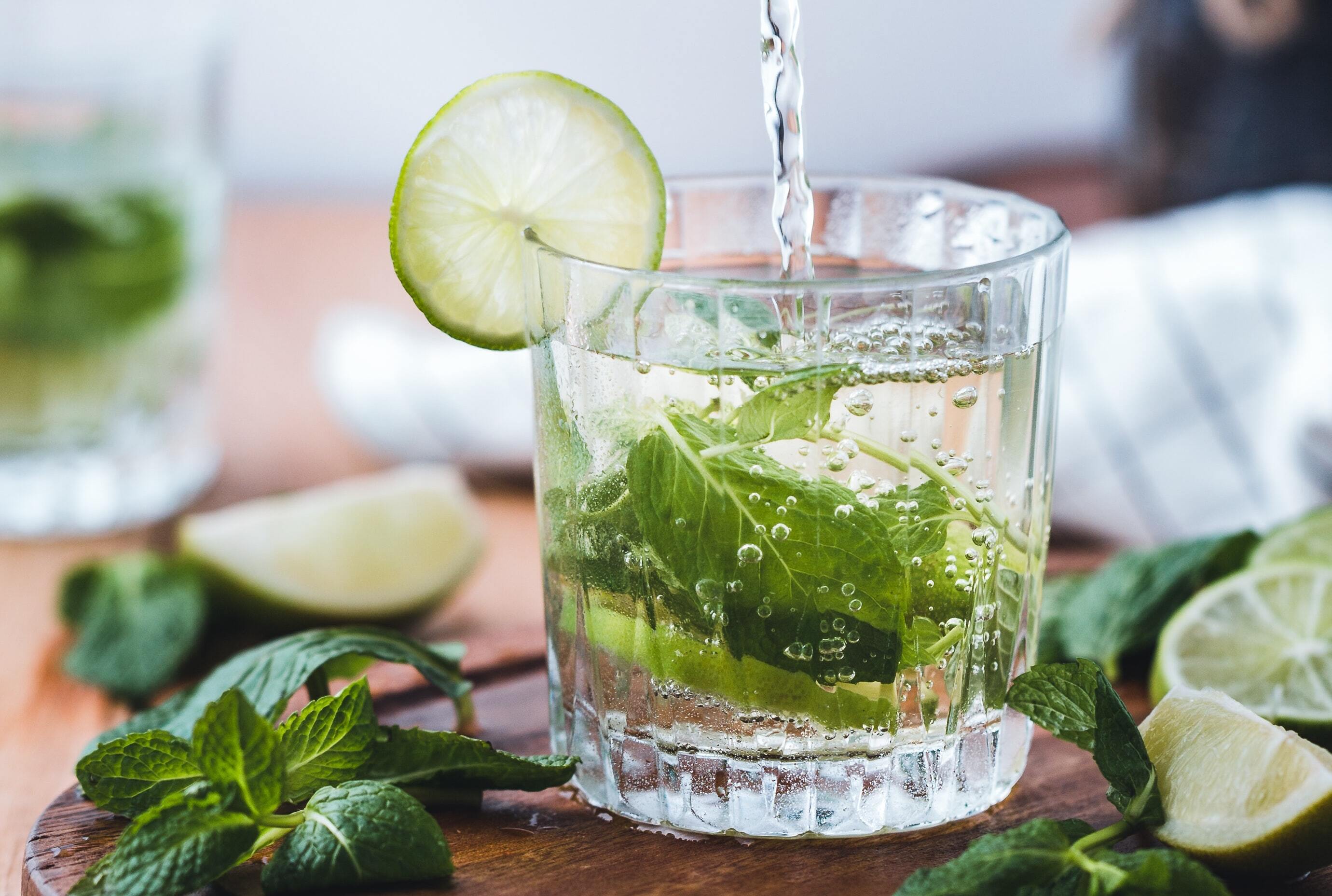 Cocktail glass being filled with a sparkling lime and mint drink, garnished with a lime slice and mint on a wooden board