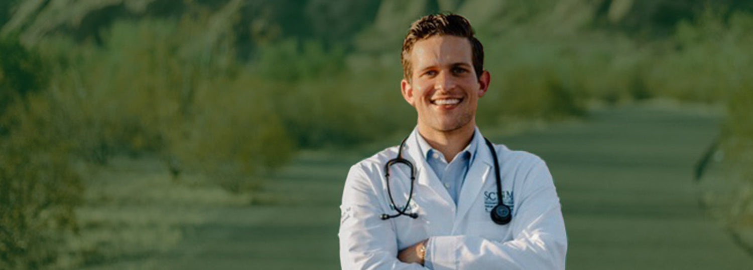 Young male doctor smiling with arms crossed, wearing a white coat and stethoscope outdoors with a blurred green background