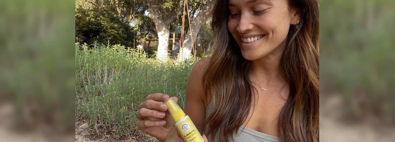 Smiling woman holding a yellow Beekeeper's Naturals propolis spray bottle outdoors in a garden