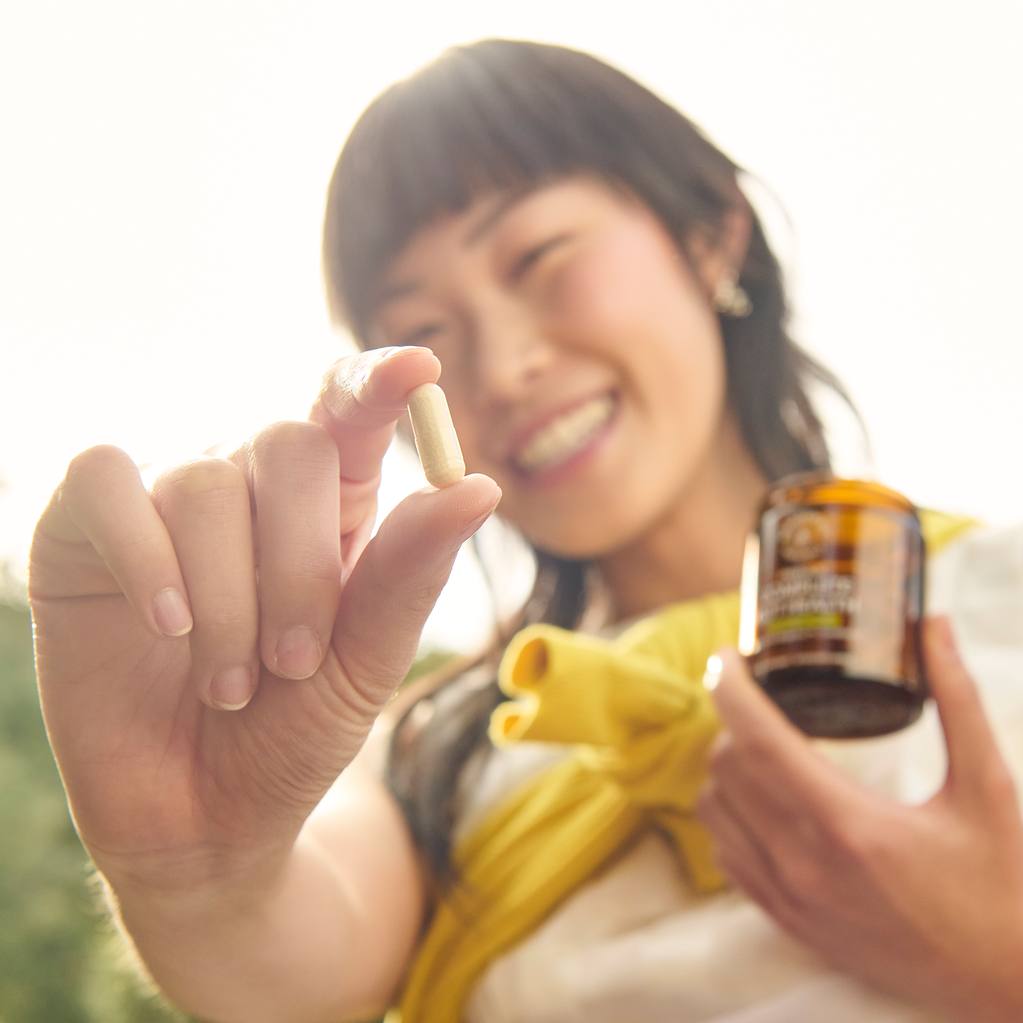 Woman smiling and holding a supplement capsule close to camera with an amber jar, bright outdoor background