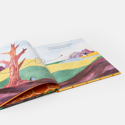 An open illustrated children's book spread on a clean white background, showing colorful landscape artwork with a tree, hills, water, and a small bird.