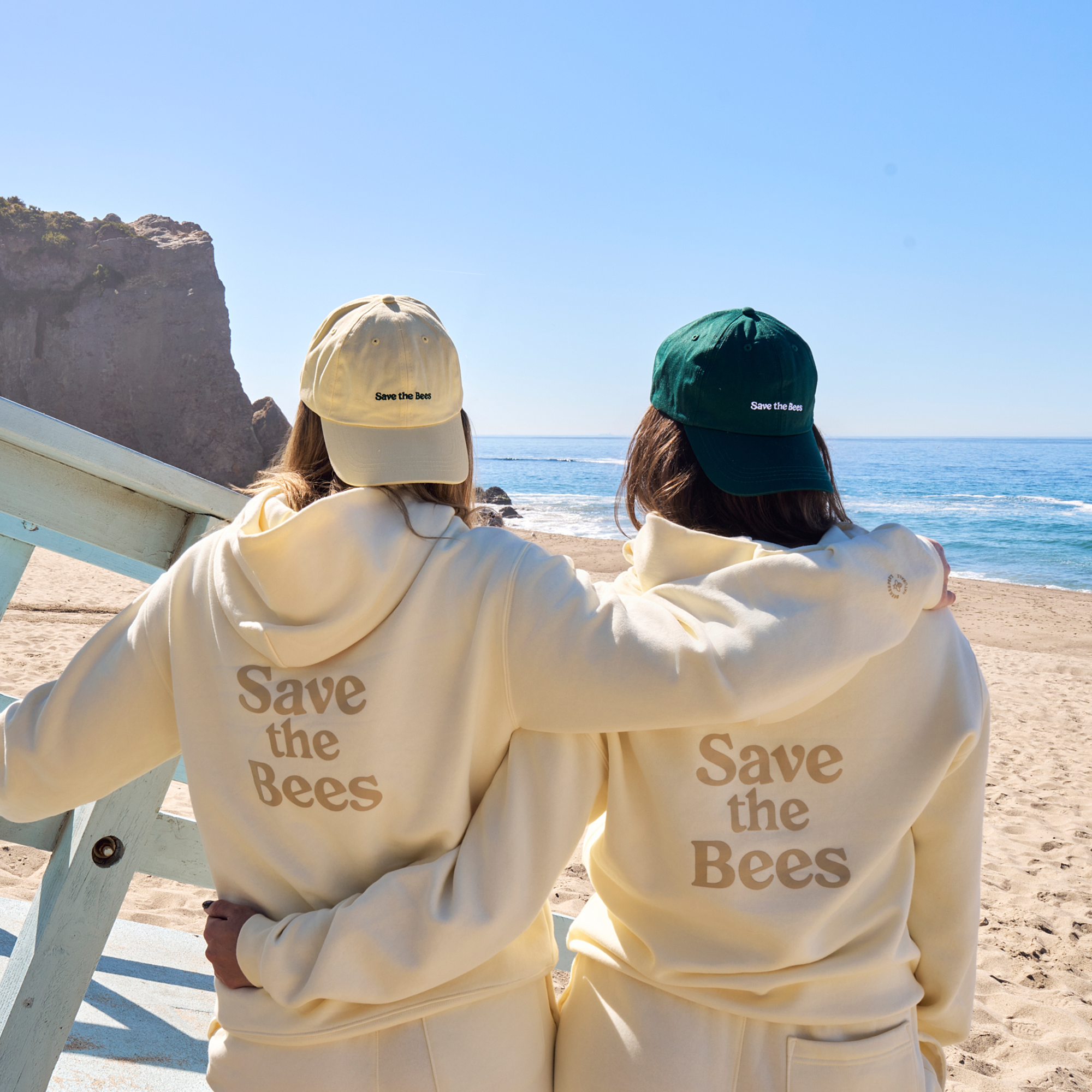 Two people on a beach wearing matching cream hoodies with 'Save the Bees' text, facing away from the camera.
