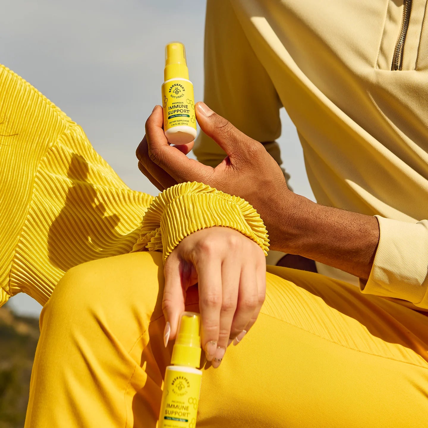 Two people holding Beekeeper’s Naturals Propolis Immune Support spray bottles outdoors.
