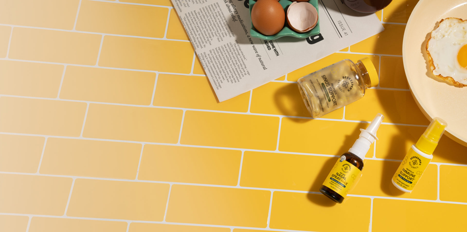 Three bottles of Beekeeper's Naturals products on a yellow tiled floor with eggs and a newspaper in the background.